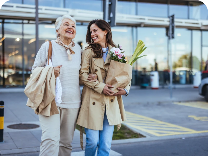 Women smiling outside, holding flowers and bags.